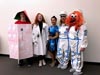 Five people lined up in a row and wearing costumes. From left to right, they are wearing: a makeshift, handdrawn-looking rocket ship, a whitecoat, a dress that has a space pattern, and two astronauts.